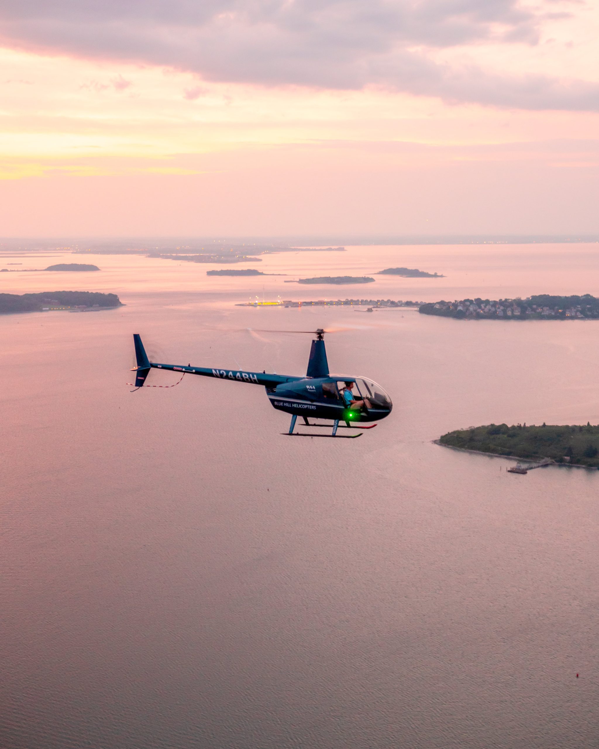 Private helicopter flying over calm coastal waters at sunset, with small islands and shoreline visible in the distance