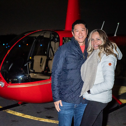 Smiling couple standing beside a red helicopter at night before a Boston helicopter tour.